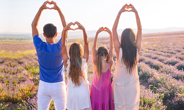 Une famille de 4 personnes, debout au milieu d’un champ de lavande. Ils se tiennent dos à l’observateur. Leurs mains sont tendues vers le haut, et montrent le symbole du cœur avec leurs doigts.