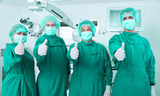 Quatre infirmières sont debout dans une salle d’opération, vêtues de vêtements stériles et montrent leurs pouces pour donner un signe positif.