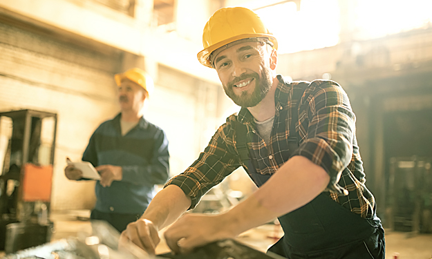 Deux artisans avec des casques se trouvent dans un bâtiment industriel. L’un d’eux déballe quelque chose mais regarde le spectateur et lui sourit.