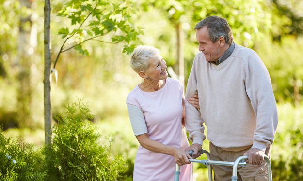 Une femme plus âgée tient son mari par le bras. Ils se promènent dans le parc de la maison de retraite et se sourient. C’est une très belle journée ensoleillée.