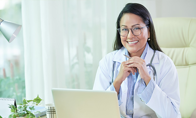 Un médecin souriant avec des lunettes, une blouse et un manteau blanc de médecin est assis à son bureau devant un ordinateur portable et regarde le spectateur avec ses beaux yeux bleus.