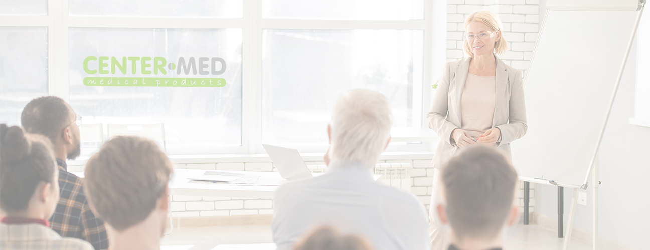 Une femme aux cheveux blonds se tient à côté d’un tableau blanc, tout en donnant une formation à un groupe d’étudiants.